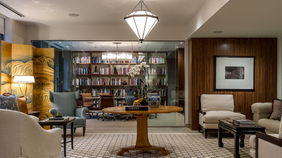 The image shows a luxurious, elegantly designed room with a blend of classic and contemporary styles. The foreground features a round wooden table topped with a decorative orchid plant, books, and blue-and-white ceramic vases. Comfortable chairs and sofas with neutral-colored upholstery are arranged around the table, and a patterned rug covers the floor. In the background, a glass-enclosed room holds bookshelves filled with books, while the walls are adorned with wood paneling and framed artwork. A large, intricate screen with gold wave patterns adds an artistic touch to the space. The overall ambiance is warm, inviting, and sophisticated.